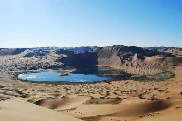 A blue lake ringed by green reeds in a basin of towering sand dunes under a clear sky in the Badain Jaran Desert, Inner Mongolia.