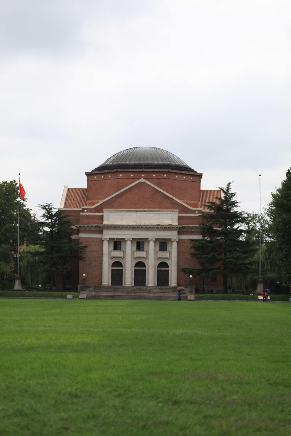 The domed red-brick Grand Auditorium of Tsinghua University in Beijing, seen across a wide green lawn under an overcast sky.