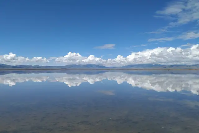 Cumulus clouds and distant mountains mirrored in the still, shallow water of Chaka Salt Lake in Qinghai under a deep blue sky.