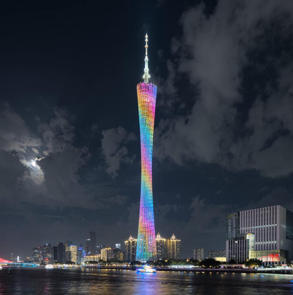 Canton Tower lit in rainbow colours beside the Pearl River in Guangzhou at night, under a cloudy sky with the moon behind the clouds.