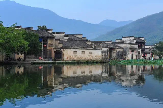 Whitewashed Huizhou houses of Hongcun village reflected in the still water of South Lake, with green mountains behind.