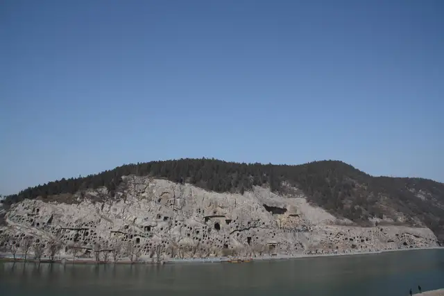 The Longmen Grottoes cliff, honeycombed with carved caves, rising above the Yi River in Luoyang under a clear blue sky