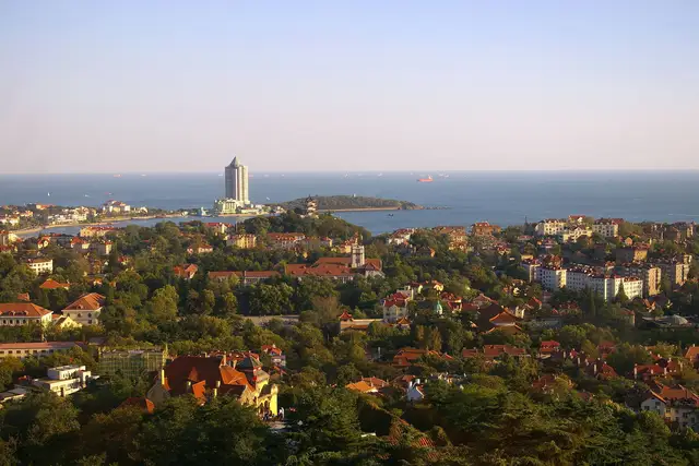 Red-tiled roofs and green trees of old Qingdao running down to the blue bay and Little Qingdao island, seen from Signal Hill in late-afternoon sun.
