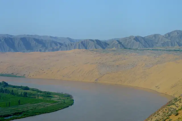 Golden sand dunes of the Tengger Desert slope straight down to a bend of the Yellow River at Shapotou, with green fields on the far bank and grey mountains beyond.