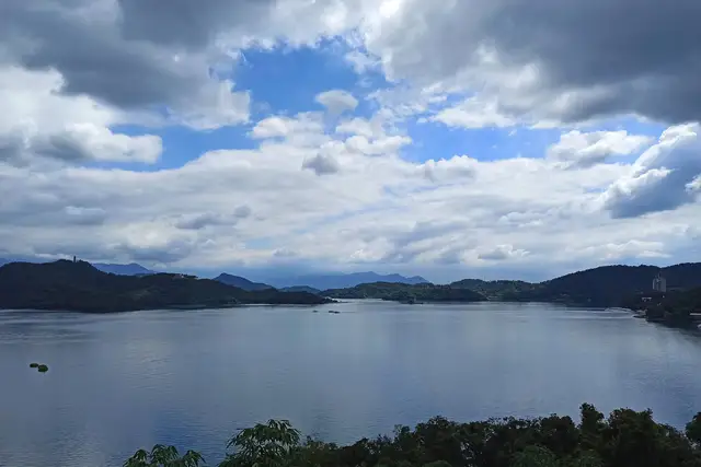 Sun Moon Lake in Nantou, Taiwan, a wide calm lake ringed by forested hills with distant mountains under a dramatic sky of white clouds and blue patches.