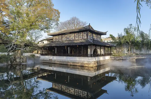 Jianshan Tower, a two-storey timber pavilion, reflected in a misty pond at the Humble Administrator's Garden in Suzhou on a clear autumn morning.