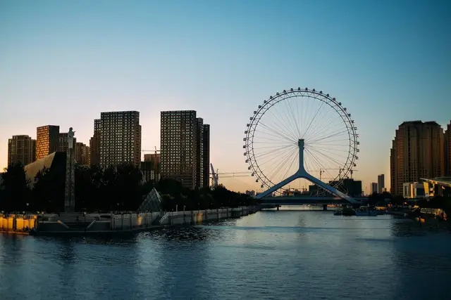 The Tianjin Eye Ferris wheel on Yongle Bridge silhouetted against a clear dusk sky above the calm Hai River, with riverside towers on both banks.