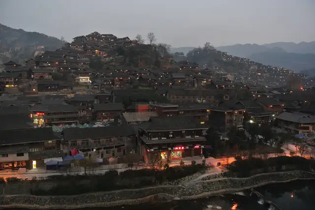 Xijiang Thousand Households Miao Village in Guizhou at dusk: dark wooden stilt houses climb the hillsides with warm lights coming on, above a riverside street and the river.