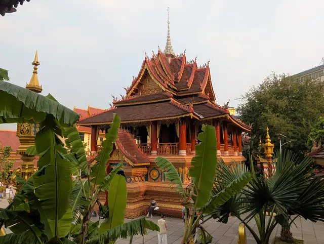 A Dai-style hall with tiered red-brown roofs and a silver spire at Zongfo Temple in Jinghong, Xishuangbanna, framed by banana and palm leaves.