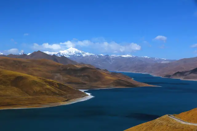 A deep-blue lake winds between golden-brown hills towards a snow-capped peak under a bright blue sky at Yamdrok Lake, Tibet.